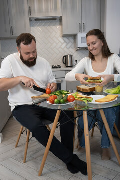 Young couple preparing a healthy meal together, slicing fresh vegetables and assembling sandwiches at the kitchen table, sharing a moment of domestic life. Vertical photo