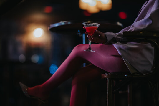 A girl sits at a bar stool holding a red cocktail in a martini glass. She wears pink tights and a white blazer in a moody, low-light bar setting.