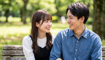 Fototapeta premium Happy young Japanese couple smiling and looking at each other while sitting on a park bench during a sunny day date in nature
