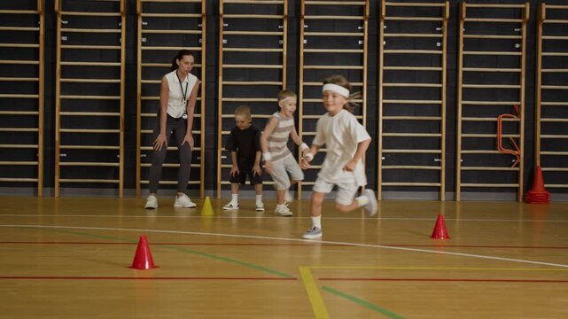 Caucasian female teacher supervising Caucasian children running relay race around cones in school gym. Clip is ideal for school sports, physical education, and youth fitness commercials
