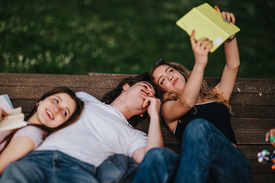 Students lie on a wooden bench in a park, taking a selfie while holding notebooks. Three students relax outdoors, sharing a casual study break and smiling at the phone.
