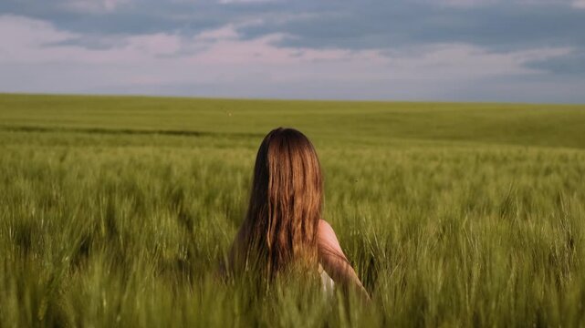 Single person with long brown hair seen from behind reaches outward into tall green wheat field while facing low horizon and rolling farmland, gentle arm extension and slight body lean create