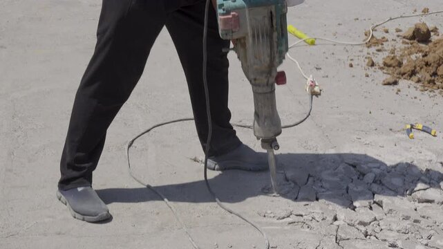 A construction worker grips an electric jackhammer with both hands and breaks cracked concrete pavement beside loose rubble during outdoor road repair.
