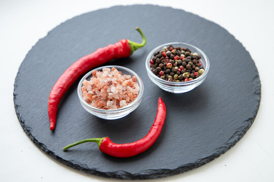 Gourmet food background closeup with organic seasoning ingredients, pink himalayan rock salt and mixed peppercorns in bowls circled by red hot chili peppers on round black slate plate.