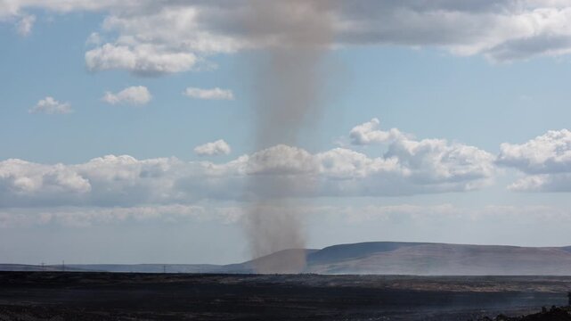Dust devil formed by intense ground heat spinning over smouldering wildfire aftermath, dark scorched earth stretching toward distant Welsh hills under summer clouds
