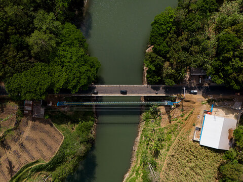Aerial view of Sengguruh Bridge spanning the river, surrounded by lush green trees, agricultural fields, and a nearby building in Malang, East Java, Indonesia.