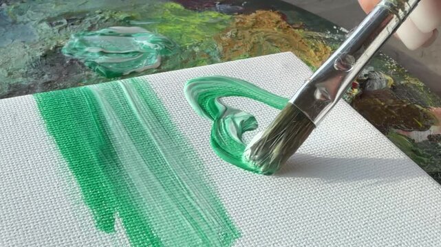 A paintbrush applies green paint to a textured canvas surface.