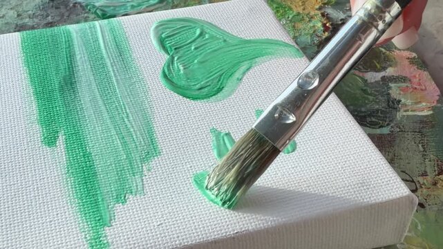 A paintbrush applying green acrylic paint on a canvas.