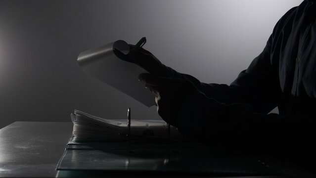 Person's silhouette sitting at a desk, carefully reviewing confidential documents within a file binder, highlighting concepts of secrecy, investigation, and private information