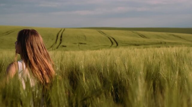 Young woman seen from behind walking slowly through tall green wheat field, then turning to face camera in partial rotation, fingertips brush stalks while light sleepless dress trails slightly