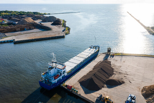 An aerial view captures a large cargo vessel docked by piles of materials at the industrial harbor of Salacgriva, Latvia, facing the sea.