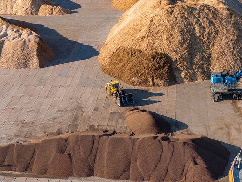 Aerial view of a yellow industrial loader working near large piles of wood chips and timber sawdust in the port storage area of Salacgriva, Latvia.