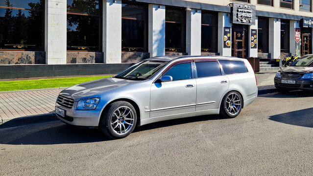 Silver Nissan Stagea M35 station wagon parked at the roadside in front of a modern building in Barnaul, a Japanese right-hand-drive modern classic, Altai Region, Siberia, Russia 25.05.25