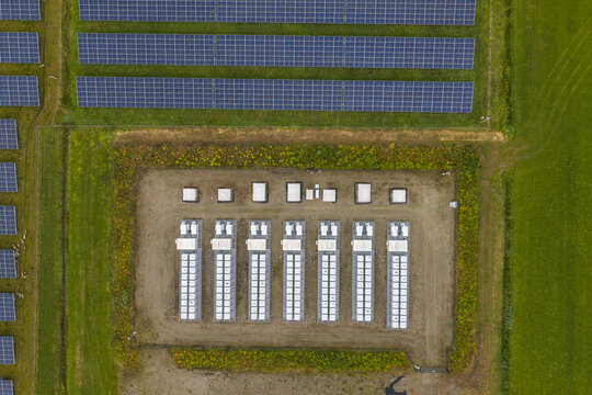 Aerial view of a solar farm featuring rows of photovoltaic panels and a central battery energy storage system with white containers on a gravel lot in the Netherlands.