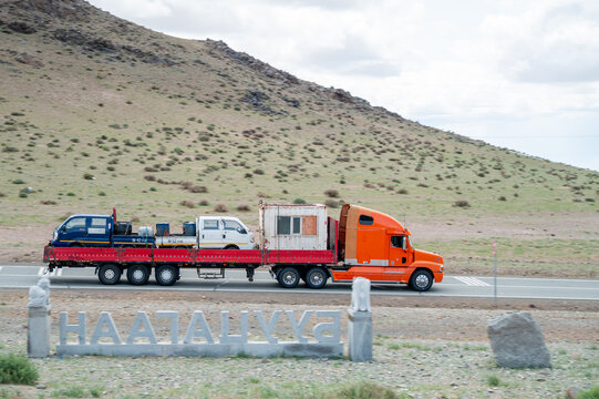 Orange heavy truck with a flatbed trailer and cargo driving through the Mongolian steppe against a backdrop of hills; freight transport on a rural road, Mongolia 02.06.2025