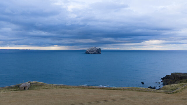 Aerial view of Bass Rock rising from the North Sea, surrounded by deep blue water under a dramatic cloudy sky, with a golden grassy field in the foreground in Scotland, United Kingdom.