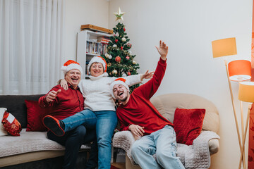 Naklejka premium A happy family of three wearing Santa hats sits together in a living room beside a decorated Christmas tree. They laugh and celebrate the holiday season on a cozy sofa in a warm home setting.