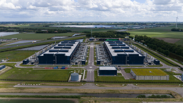 Aerial view of a large modern data center facility with two massive industrial buildings, rooftop cooling units, and surrounding green fields in Middenmeer, North Holland, Netherlands.