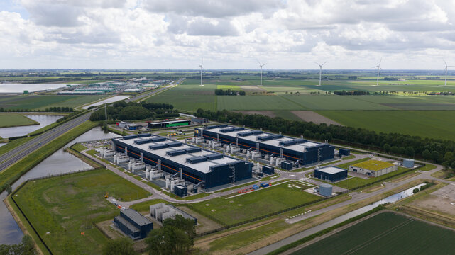 Aerial view of a large data center facility surrounded by green agricultural fields, wind turbines, and canals in Middenmeer, North Holland, Netherlands.