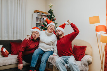 Naklejka premium A group of family friends wearing Santa hats sit on a couch by a decorated Christmas tree. They pose playfully together in a cozy living room holiday scene.