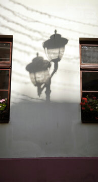 Shadow of double street lamp cast on light wall between two windows with flower boxes. Symmetry of architecture and contrast of sunlight create artistic urban composition