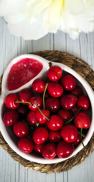 White ceramic cherry bowl with separate pit section. Bright red cherries with stems on woven mat, rustic summer food composition highlighting freshness and practical design