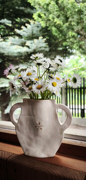 White daisies with yellow centers in handmade ceramic vase on a wooden windowsill. Soft light and garden view create a peaceful, natural decorative scene
