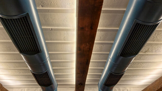 Exposed blue grey industrial air ducts with vents running parallel on a white textured ceiling, separated by a rustic wooden beam, creating a modern, functional aesthetic