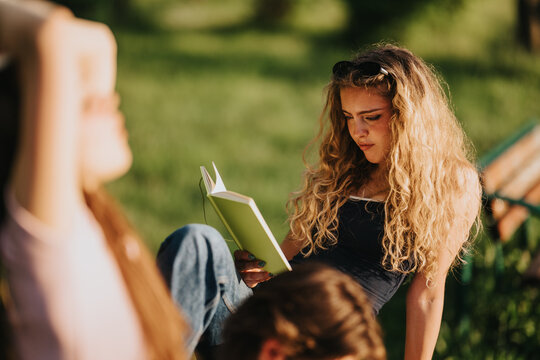 A teenage girl reads a book while sitting outdoors in a sunlit park. She relaxes on the grass and concentrates on her reading.