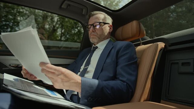Middle aged Caucasian businessman sitting in back seat of moving luxury car, analyzing financial documents. Clip is suitable for corporate travel, executive lifestyle, or wealth management ads