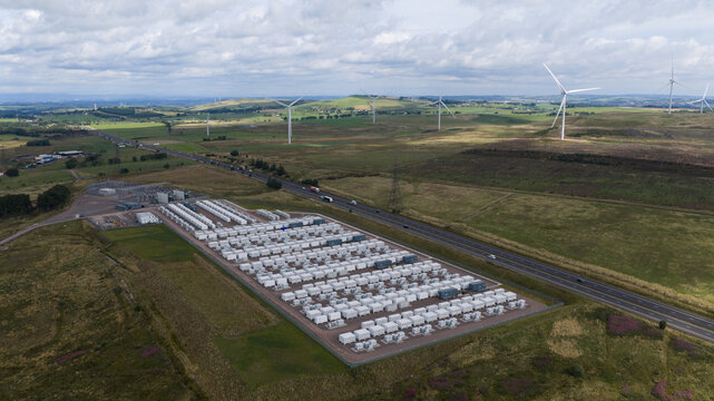 Aerial view of the Coalburn BESS facility featuring rows of white battery storage containers set within a rural landscape with wind turbines and a highway in Lanark, Scotland, United Kingdom.