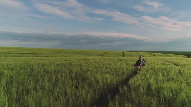Two people walk between tall green cereal stalks along narrow tractor tracks across rolling farmland, one wearing wide brim hat and denim jacket, with open horizon, soft light, and peaceful rural