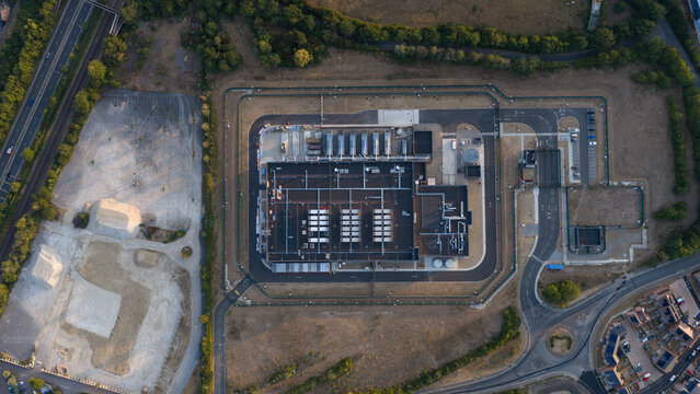 Aerial view of a modern data center facility with rooftop cooling units, surrounded by a secure fence, access roads, and nearby residential housing in Bracknell, England, United Kingdom.