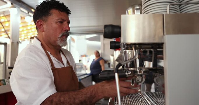 Professional middle aged barista with a goatee using a chrome coffee machine to pour a fresh hot espresso into a white cup inside a busy modern cafe or coffee shop
