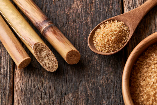 Raw brown sugar in a wooden spoon and bowl sits beside cut sugarcane stalks, rustic food, natural sweetener, baking ingredient.