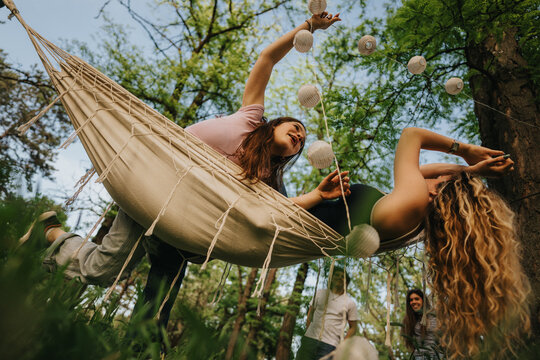 Teen friends are lounging together in a hammock decorated with paper lanterns. They relax and laugh outdoors in a wooded park setting.