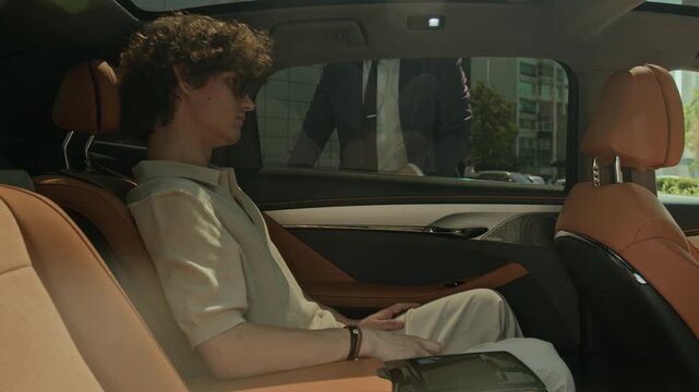 Caucasian young adult man sitting in back seat of luxury car, waiting for chauffeur to open door, then exiting vehicle. Footage suits luxury transport or executive travel promotions
