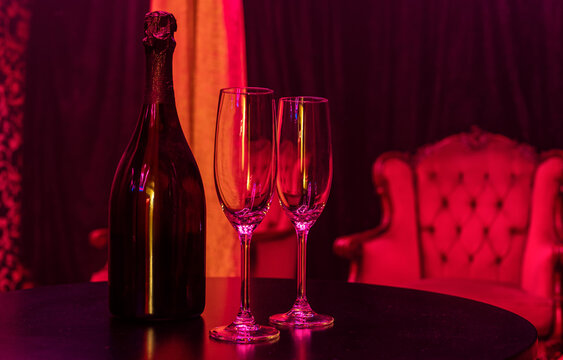 Champagne bottle and two empty flutes in romantic red light lounge.