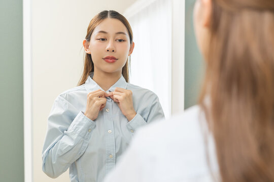 Get dress, asian young woman, businesswoman standing, hand buttoning up shirt formal, female getting dressed, ready before go to work looking reflection the mirror in bedroom in the morning at home.