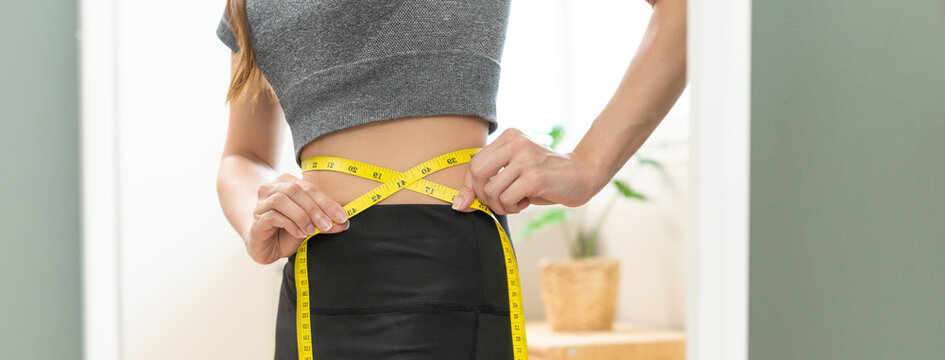 Weight lose, loss concept. Slim body asian young woman hand use tape measuring around waistline in fit sports. Girl looking reflect in mirror at home. Healthy nutrition, fitness for wellbeing beauty.