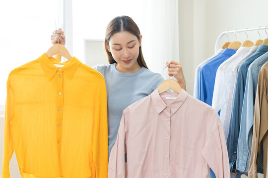 Choose clothes to wear, attractive asian young woman, girl try on appare, happy choice of shirt, female outfit on hanger in wardrobe in room closet at home. Deciding blouse what to put on which one