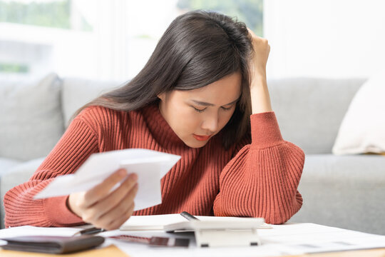 Business financial concept, stressed asian young woman hand checking list with bill for calculate accounting expense credit card payment on due deadline, trying to find money to pay debt on table.