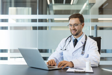 Fototapeta premium Male doctor wearing a lab coat and stethoscope, smiling while using a laptop for an online medical consultation, symbolizing remote healthcare and digital communication