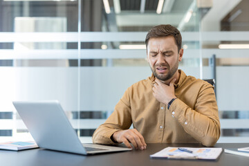 Obraz premium Young man working in an office holding his neck due to severe sore throat pain, representing illness, discomfort, and health issues impacting productivity