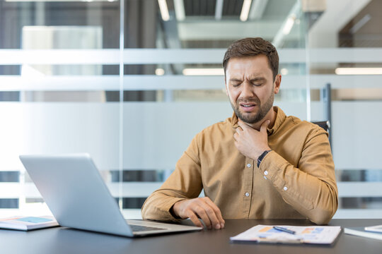 Young man working in an office holding his neck due to severe sore throat pain, representing illness, discomfort, and health issues impacting productivity