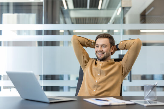 Smiling businessman leaning back in his office chair with hands behind head, enjoying a moment of relaxation and satisfaction after achieving goals and completing tasks