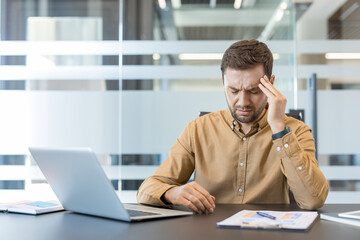 Fototapeta premium Businessman feeling severe headache and pain from overwork and stress, sitting at a desk with a laptop and documents in a modern office, experiencing professional burnout and frustration