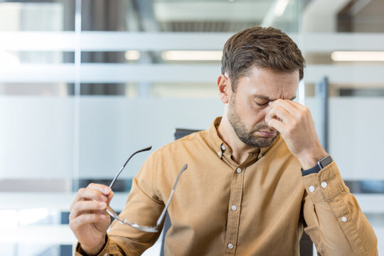 Stressed businessman rubbing his temples, experiencing a painful headache and eye strain after long hours of work, showing signs of fatigue and burnout in an office environment