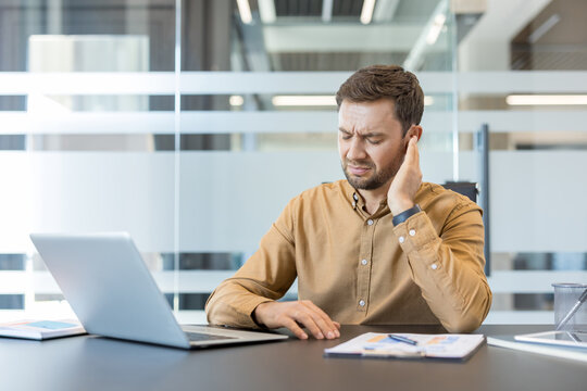 Young man experiencing discomfort and pain in his ear while working on a laptop at his office desk, indicating hearing problems, ear infection, or workplace stress affecting his health