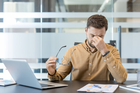 Overworked businessman experiencing burnout and fatigue, rubbing his eyes from headache and prolonged screen time while sitting at his desk with a laptop and documents